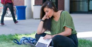 Student sitting on grassy patch of campus working on laptop.