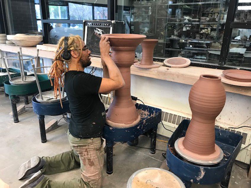 Student kneeling on floor of ceramics studio working on a large (2 feet 6 inches wheel thrown form). There is a second large wheel thrown form on the adjacent potters wheel.