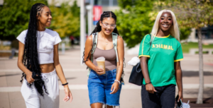 Three students walk together, smiling and laughing, outside on the MSU Denver campus en español: Tres estudiantes caminan juntos, sonriendo y riendo, en el campus de MSU Denver.